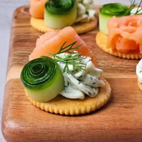 Smoked salmon cracker bites topped with whipped lemon dill cream cheese, cucumber ribbons, and fresh dill on a wooden serving board
