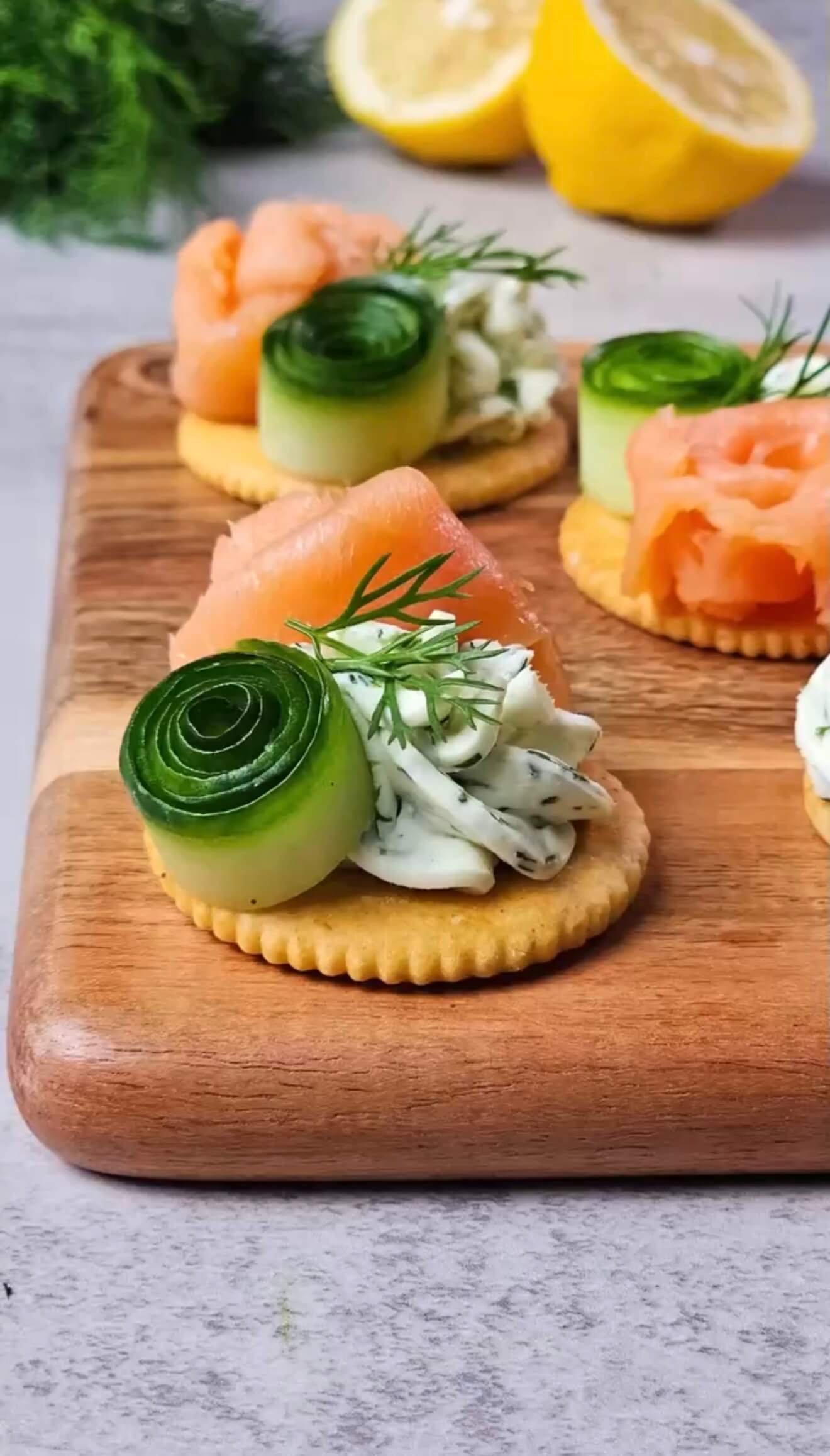 Smoked salmon cracker bites topped with whipped lemon dill cream cheese, cucumber ribbons, and fresh dill on a wooden serving board