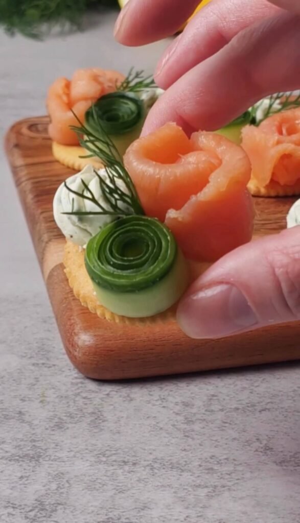 Assembling smoked salmon cracker bites with whipped herb cream cheese, cucumber roll, and folded smoked salmon