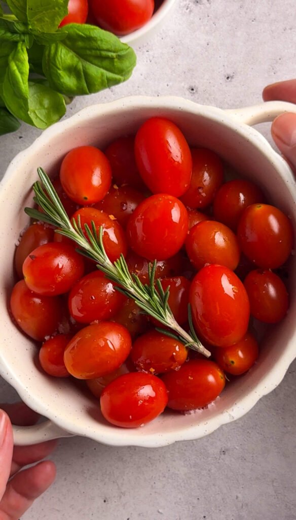 Fresh cherry tomatoes tossed with olive oil, salt, and a rosemary sprig in a small white bowl, ready for roasting.