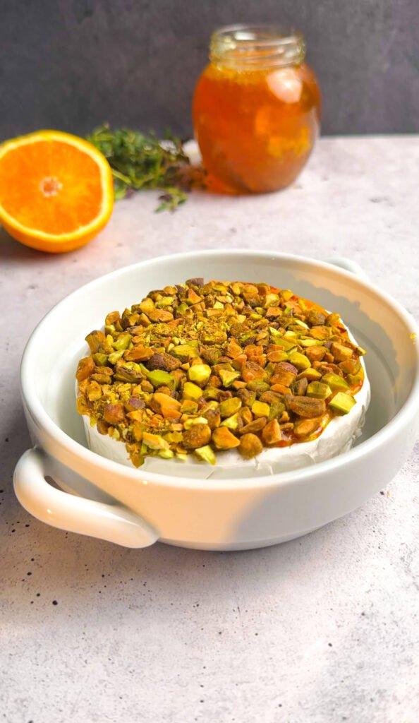 Unbaked brie wheel topped with chopped pistachios in a baking dish with honey jar and fresh orange in background.