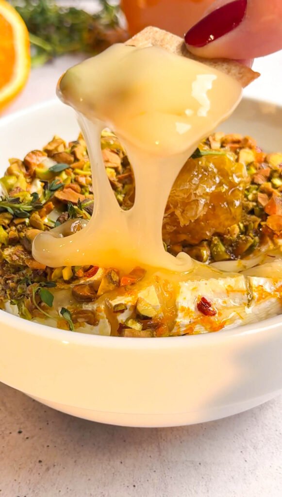 Melted pistachio baked brie with honeycomb and toasted pistachio crust stretching as it’s lifted from the dish.