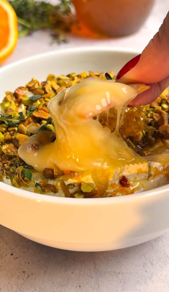 Creamy oven baked brie with honey and pistachios being scooped with a cracker, showing gooey melted texture.