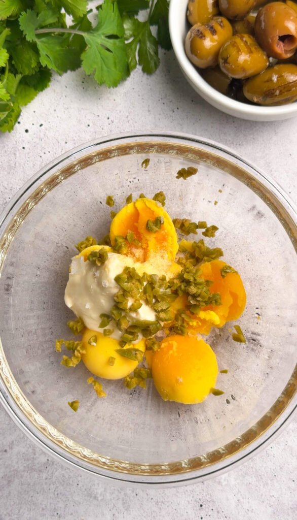 Dirty martini deviled egg filling ingredients in a glass bowl with egg yolks, mayonnaise, chopped green olives, and olive brine