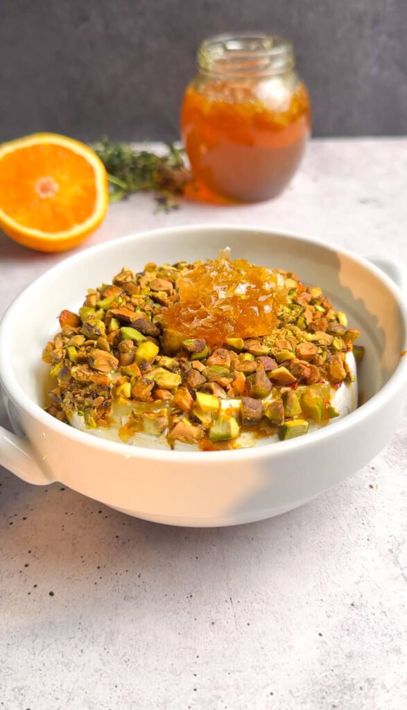 Baked brie appetizer finished with honeycomb, pistachios, and thyme in a white ceramic dish.