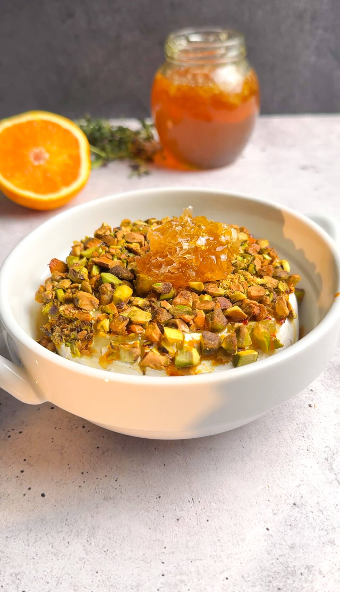 Baked brie appetizer finished with honeycomb, pistachios, and thyme in a white ceramic dish.