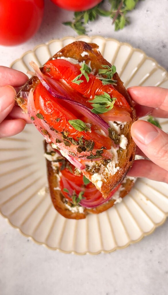 Roasted tomato feta toast with honeyed red onions and fresh oregano on sourdough, held in hands