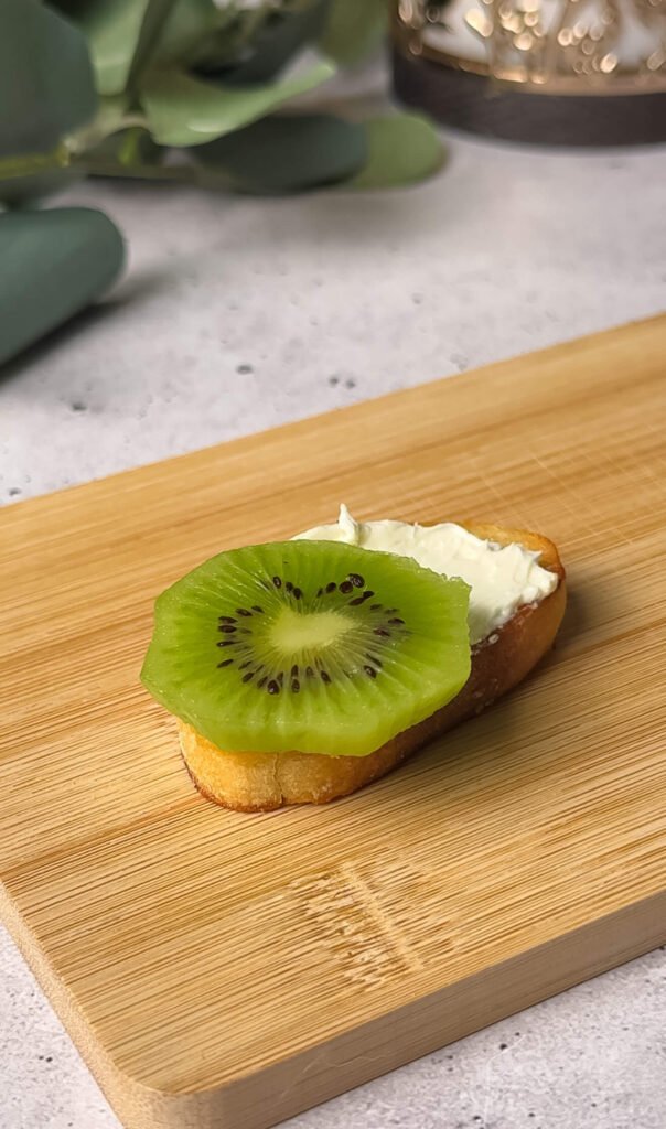 Preparing kiwi prosciutto crostini with cream cheese spread and fresh kiwi slice on toasted baguette