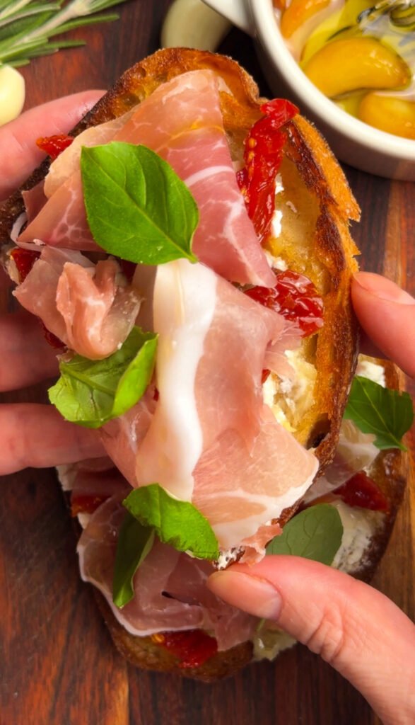 Prosciutto goat cheese toast with garlic confit, sun-dried tomatoes, honey, and fresh basil on crispy sourdough bread