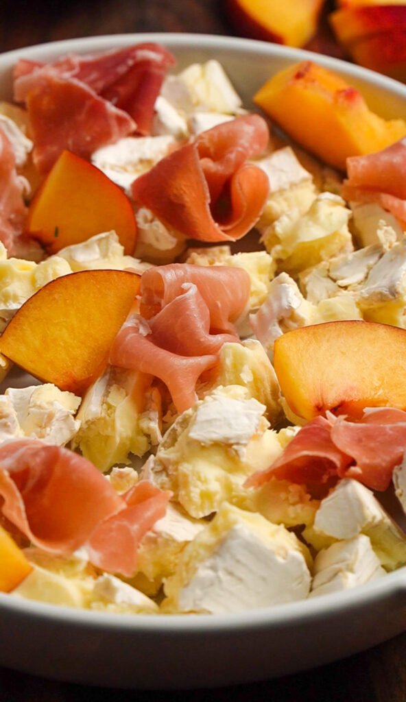 Baked brie with peach slices and prosciutto layered over soft cheese before baking