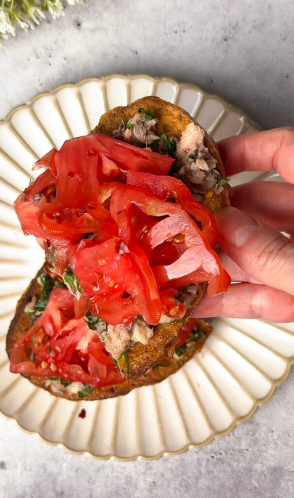hand holding heirloom tomato toast with sardines and fresh herbs