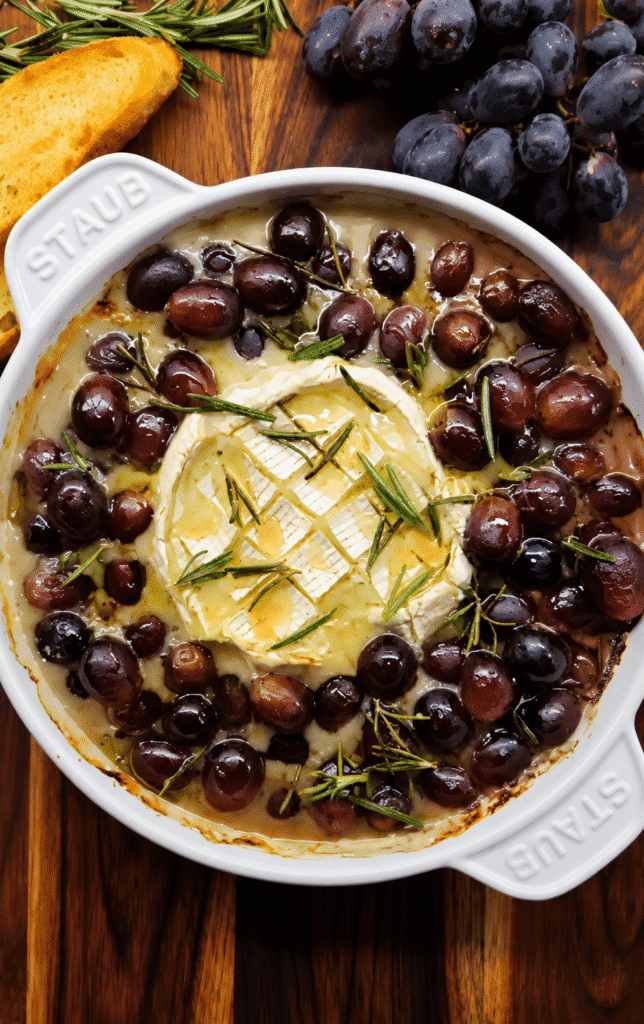 A white Staub baking dish filled with creamy baked brie topped with rosemary and olive oil, surrounded by roasted red grapes on a rustic wooden surface, served with toasted bread slices.