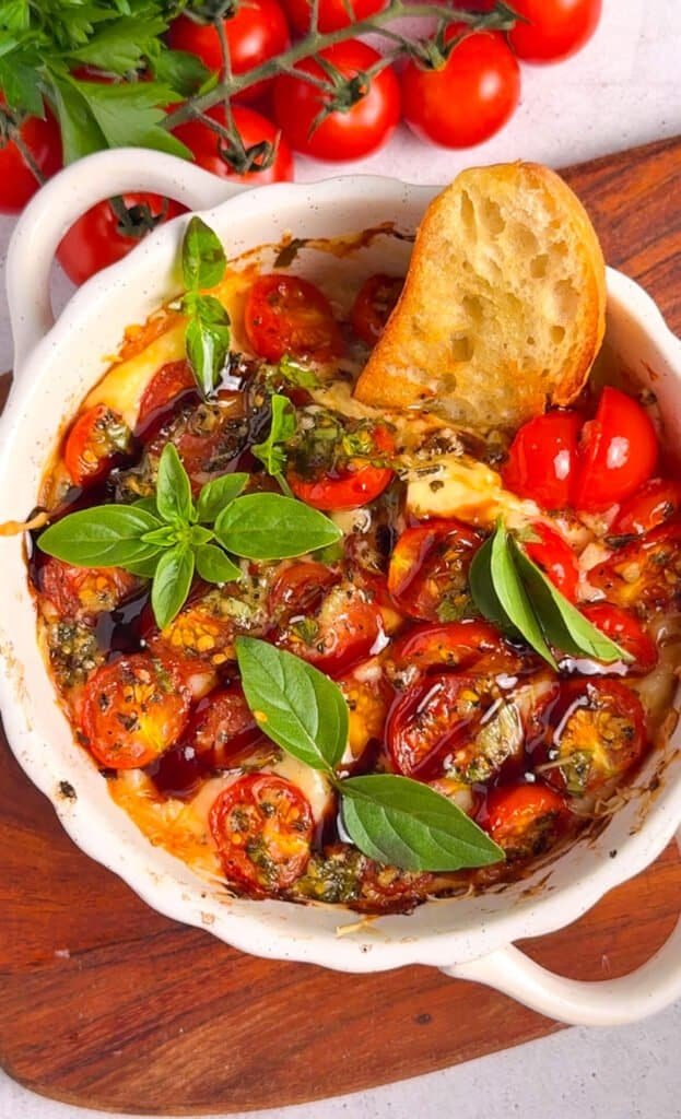 Baked tomato bruschetta dip with caramelized cherry tomatoes, fresh basil, and a golden crostini resting in the dish, served on a wooden board