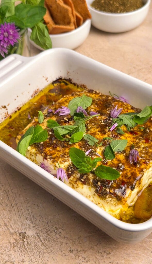 Finished baked feta with za'atar honey, fresh oregano leaves, and purple chive blossom florets in a white ceramic dish