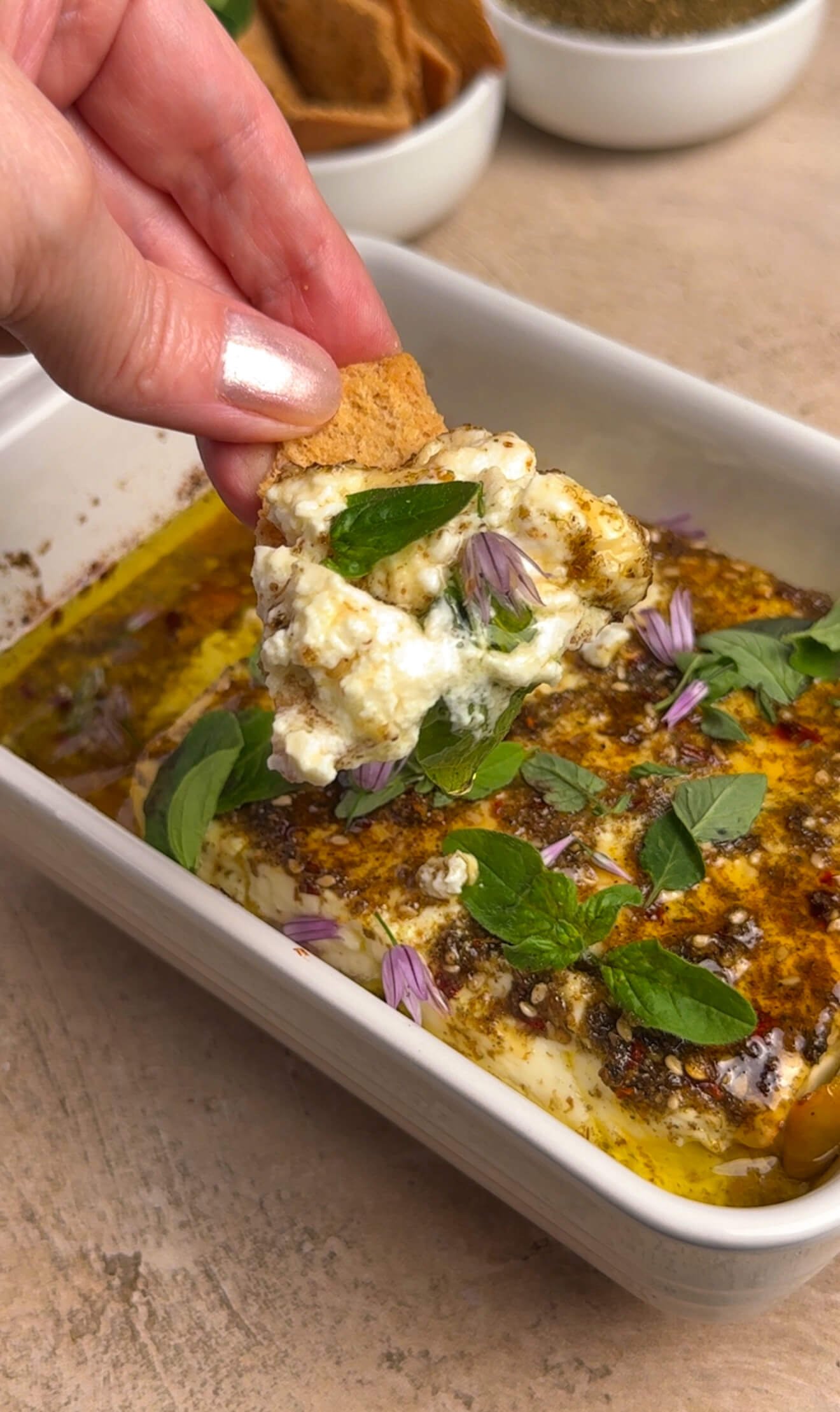 Close-up of a pita chip being pulled through warm za'atar honey baked feta with oregano and chive blossoms scattered on top
