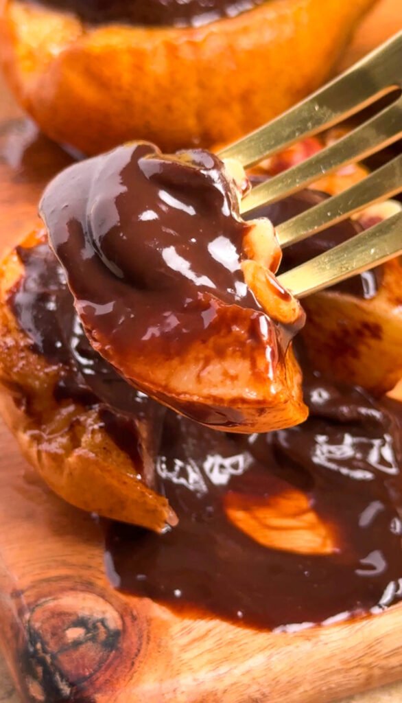 A gold fork lifting a piece of baked pear coated in rich dark chocolate ganache sauce, showing the glossy texture and caramelized fruit underneath