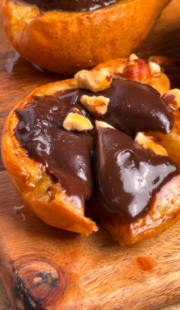 A baked cinnamon pear half sliced open and filled with dark chocolate ganache, topped with chopped hazelnuts, on a wooden board — a simple elegant fall dessert recipe