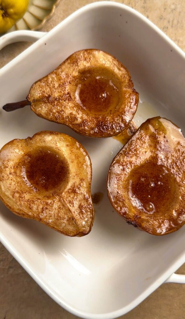 Three caramelized baked pear halves in a white ceramic baking dish, golden brown and sticky with honey after roasting