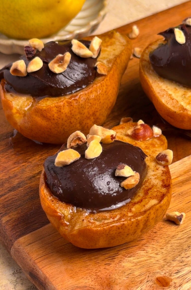 Baked pear halves filled with glossy dark chocolate ganache and topped with chopped hazelnuts, served on a rustic wooden board with a fresh pear in the background