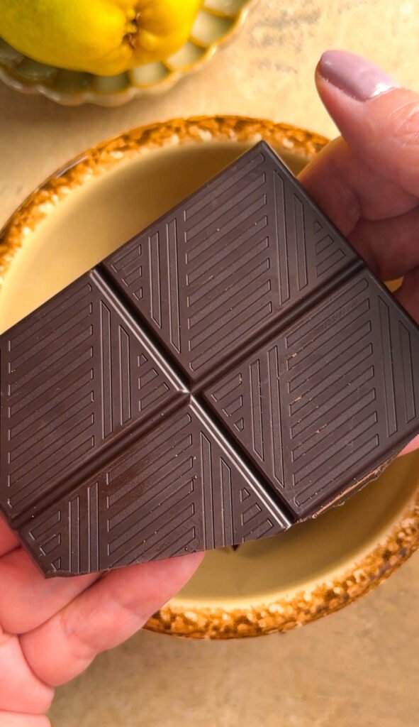 Hands holding a dark chocolate bar over a ceramic plate, with fresh pears in the background, ingredients for baked pears with chocolate sauce