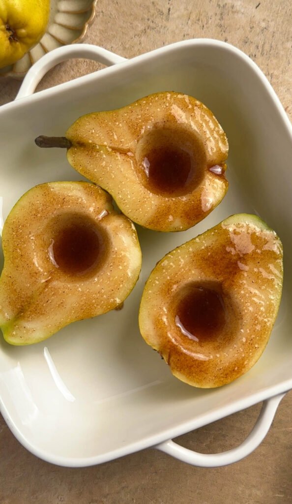 Three fresh pear halves arranged in a white baking dish, drizzled with honey and dusted with cinnamon, ready to go into the oven