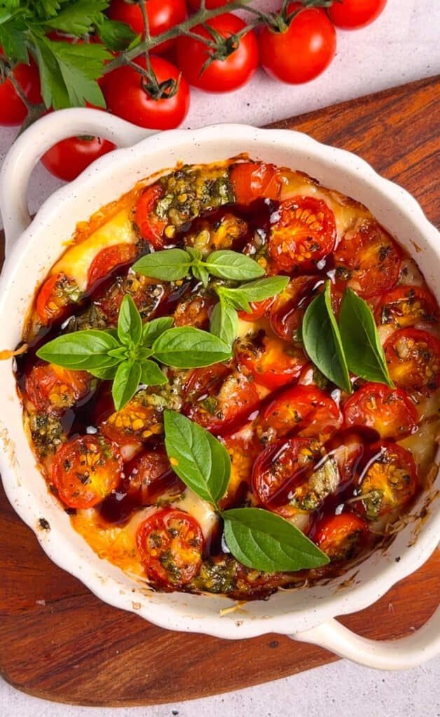 Overhead view of baked tomato bruschetta dip in a white ceramic dish, finished with balsamic glaze and fresh basil leaves
