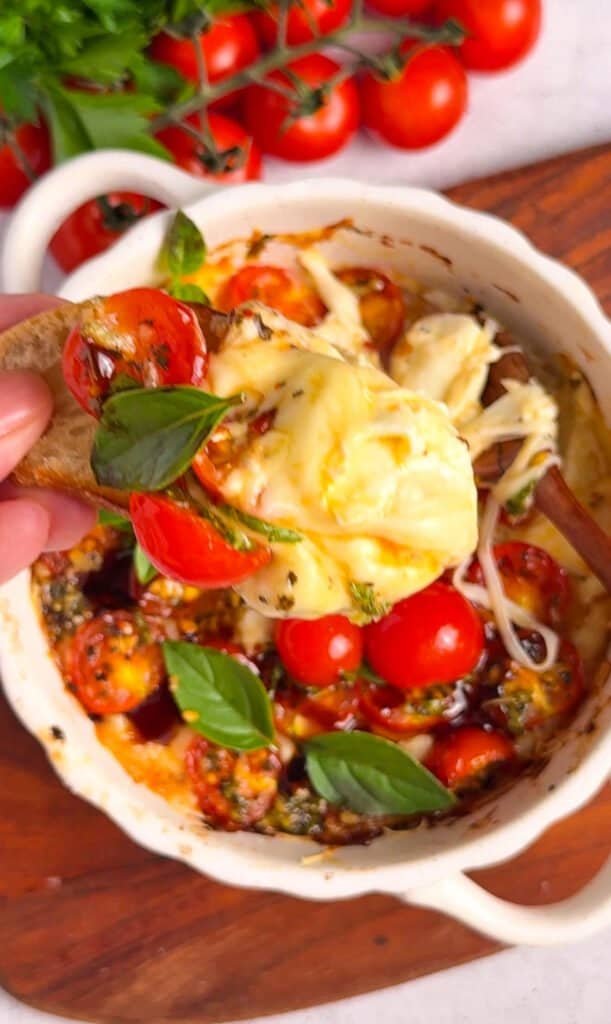 Hand scooping warm baked bruschetta dip with a cracker, showing the melted cheese pull and jammy roasted tomatoes