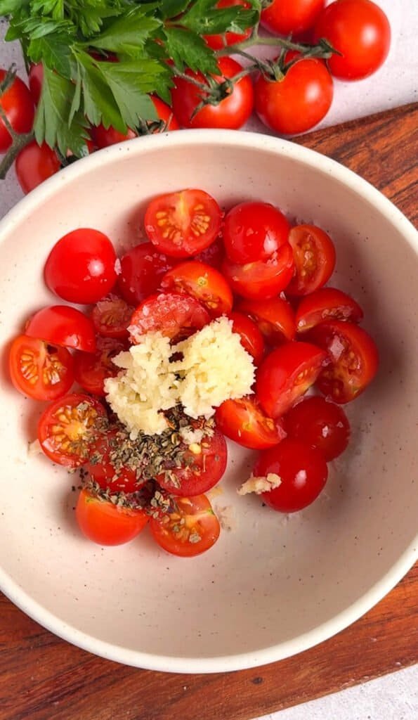 Halved cherry tomatoes with grated garlic and dried oregano in a ceramic bowl, ready to mix for baked bruschetta dip