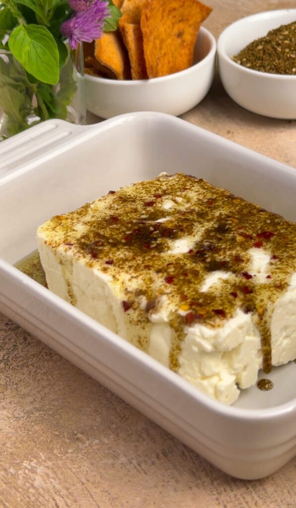 Block of feta in a white baking dish coated in za'atar olive oil and chili flakes, ready to go in the oven