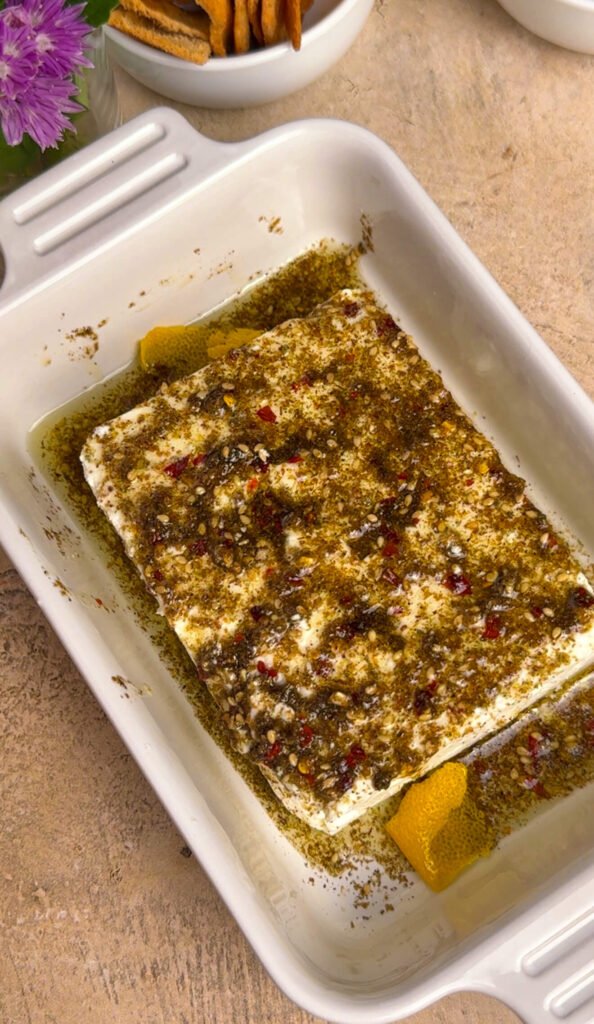 Overhead view of feta block covered in za'atar spice oil with lemon peel tucked alongside in a white ceramic baking dish