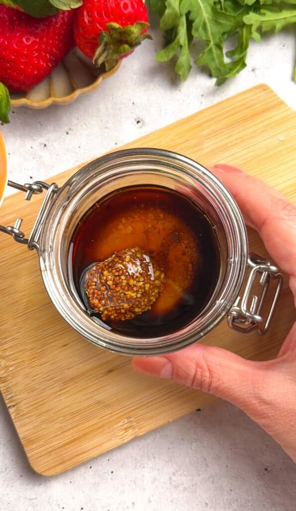 Honey balsamic dressing with whole grain mustard in a glass jar, with fresh strawberries and arugula in the background