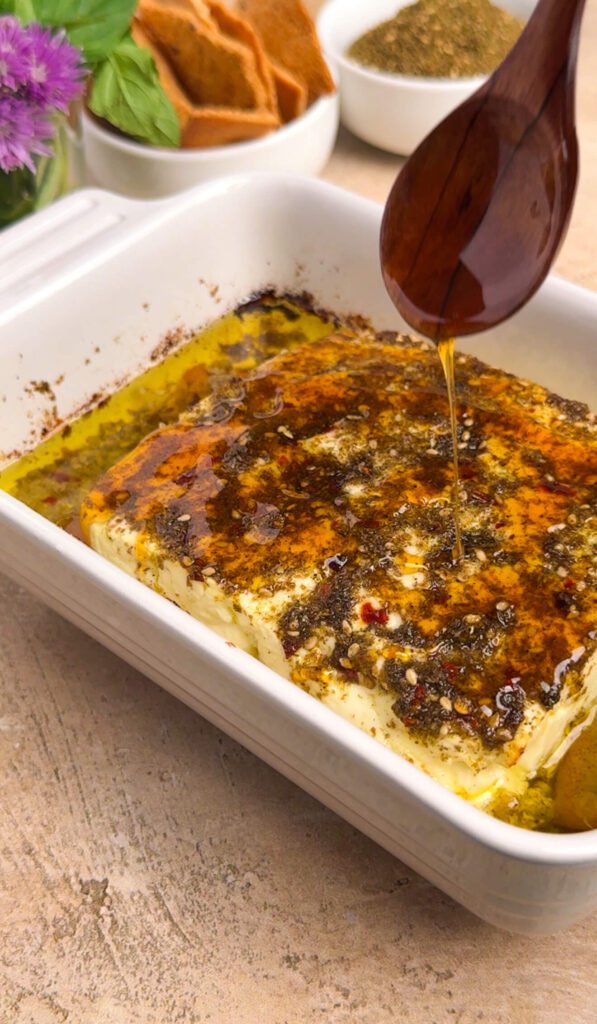 Honey being drizzled from a wooden spoon over warm baked feta with za'atar and chili oil, fresh from the oven