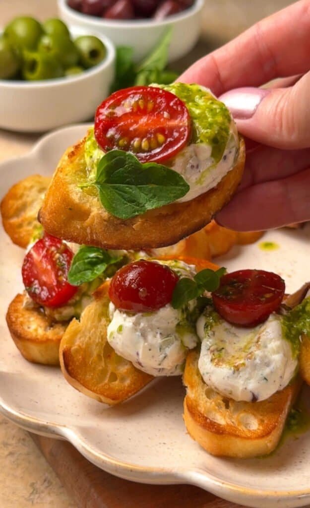 Hand picking up a whipped labneh crostini topped with cherry tomatoes, basil pesto drizzle, and fresh oregano leaves