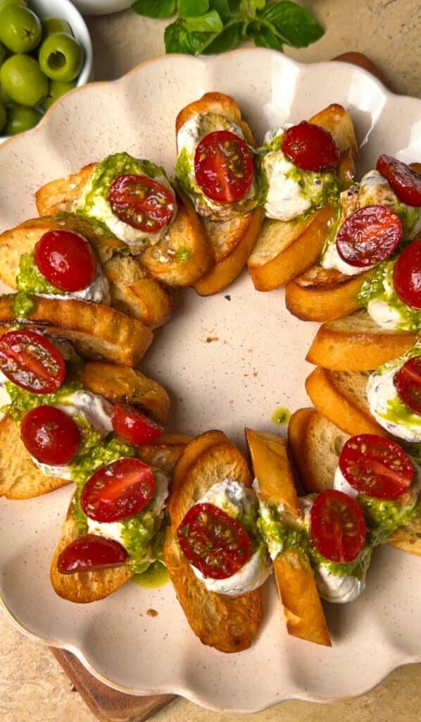 Overhead view of labneh and feta crostini arranged in a wreath shape on a speckled ceramic plate with cherry tomatoes and pesto drizzle