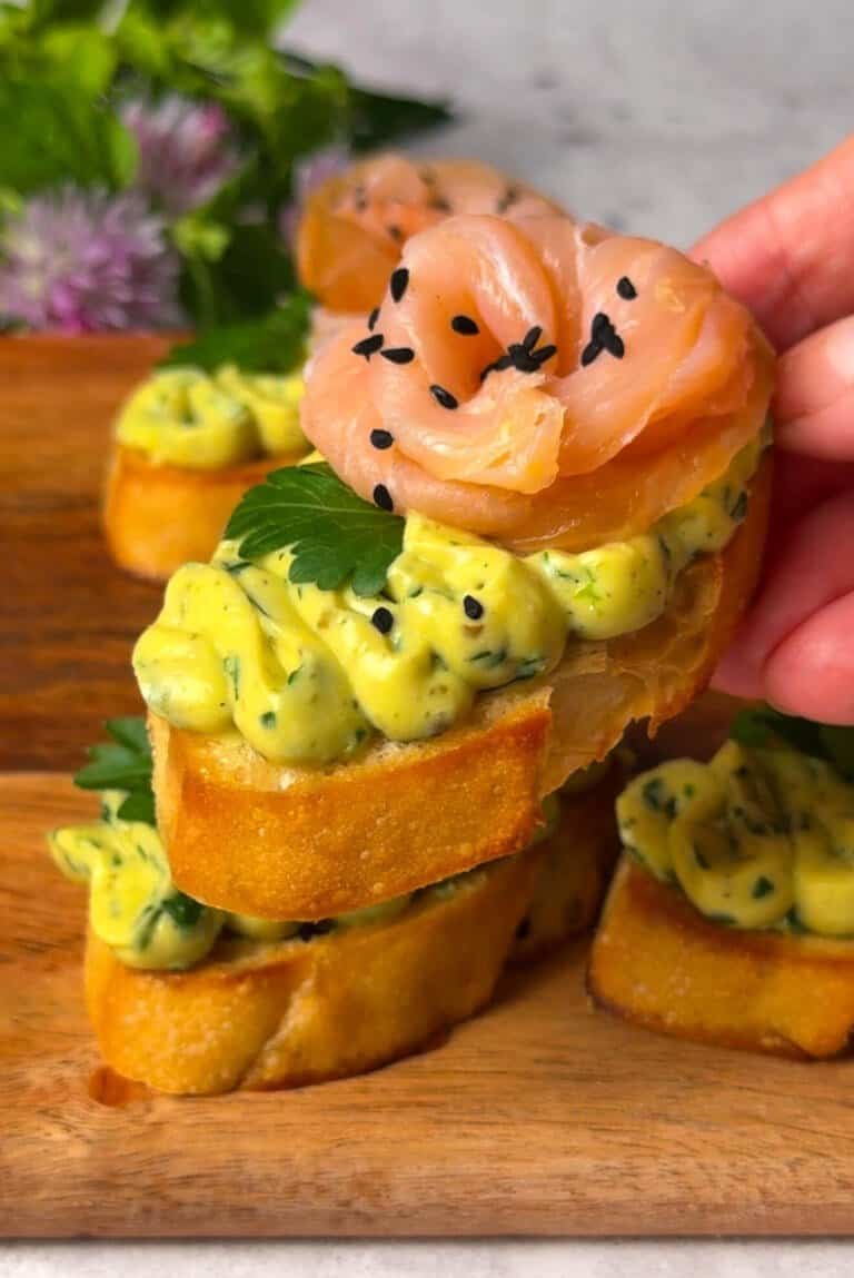 Hand holding a lemon-caper smoked salmon crostini with whipped egg spread, salmon rose, parsley, and nigella seeds