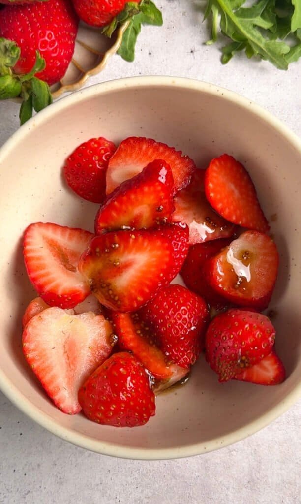 Sliced fresh strawberries macerating in honey balsamic dressing in a ceramic bowl, releasing their juices