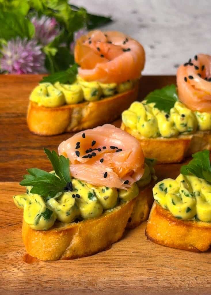 Three smoked salmon crostini on a wooden board topped with whipped egg spread, salmon roses, parsley, and black nigella seeds