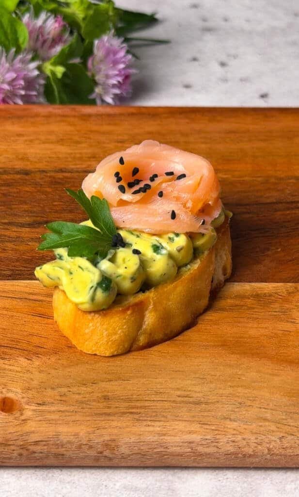 One finished smoked salmon crostini with a salmon rose, whipped green spread, parsley, and nigella seeds