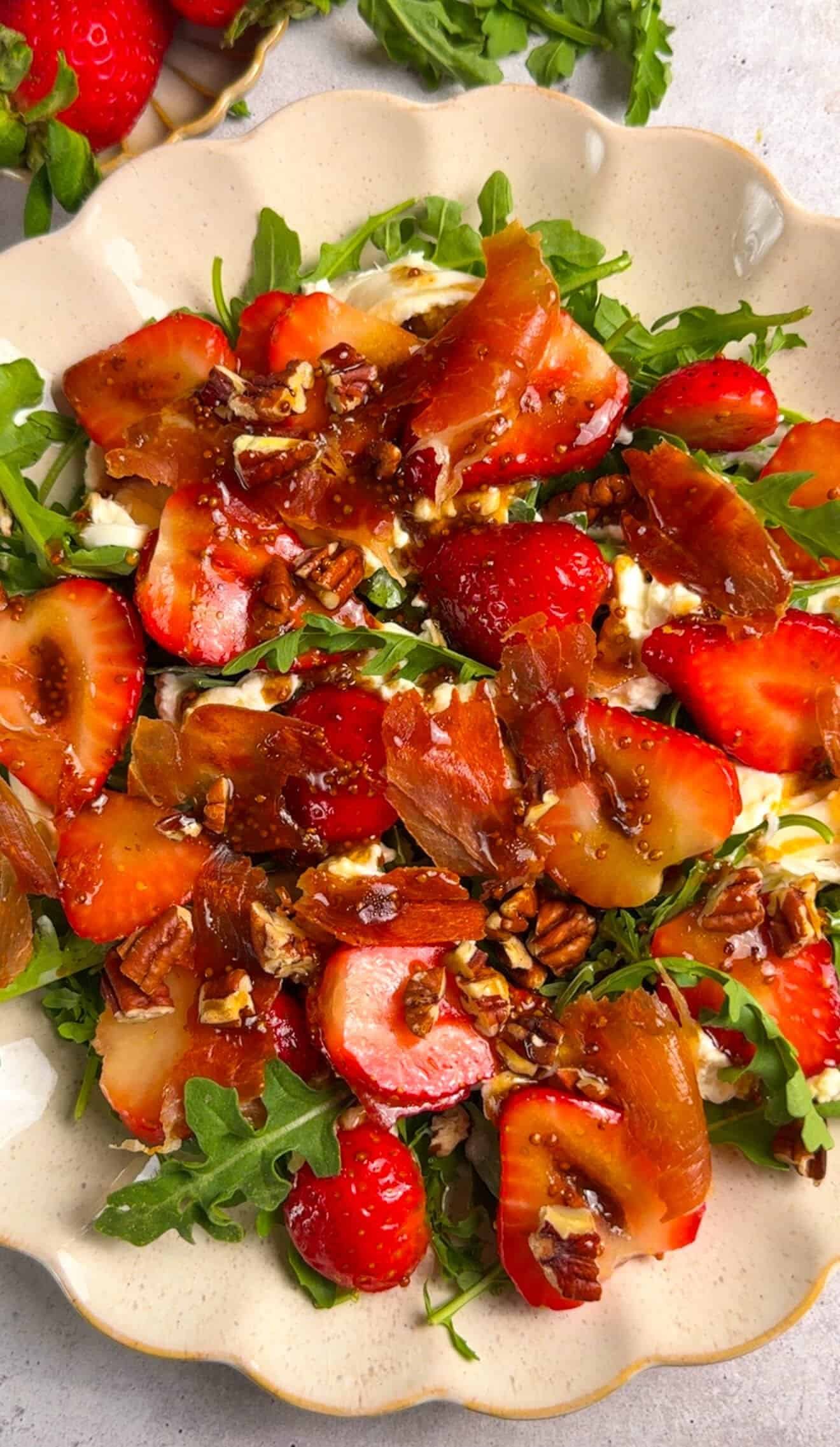 Fully assembled strawberry arugula salad with burrata, crispy prosciutto, toasted pecans, and honey balsamic dressing on a scalloped platter