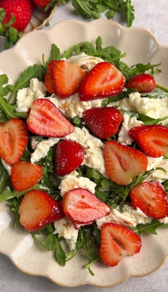 Strawberry arugula salad with torn burrata on a scalloped ceramic platter, overhead close-up view