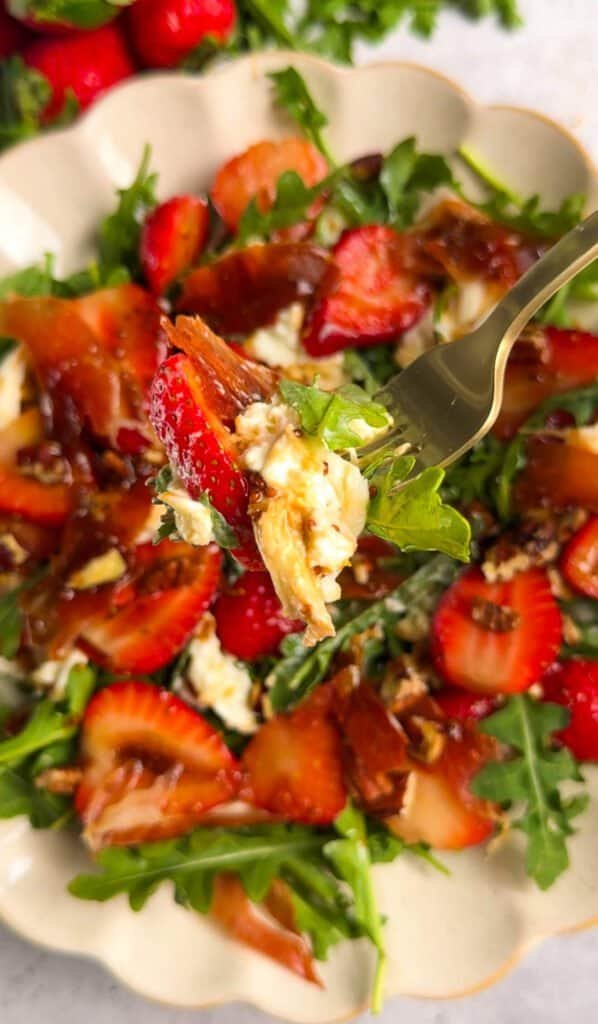 Gold fork lifting a bite of strawberry arugula salad with burrata and crispy prosciutto, showing all the layers of the dish