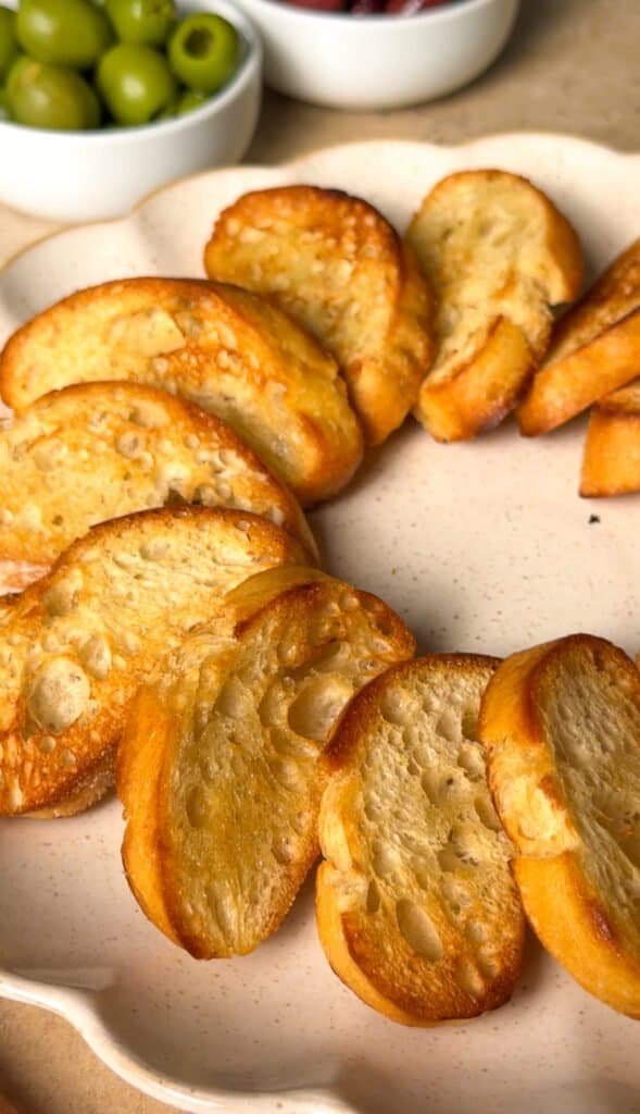 Golden toasted baguette slices arranged in a wreath shape on a scalloped ceramic plate, ready to be topped with labneh mixture