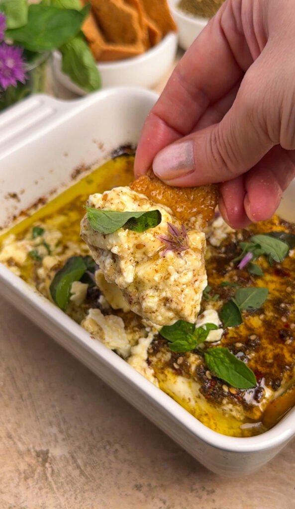Hand scooping warm baked feta with a pita chip, showing the soft creamy texture with chive blossoms and oregano on top