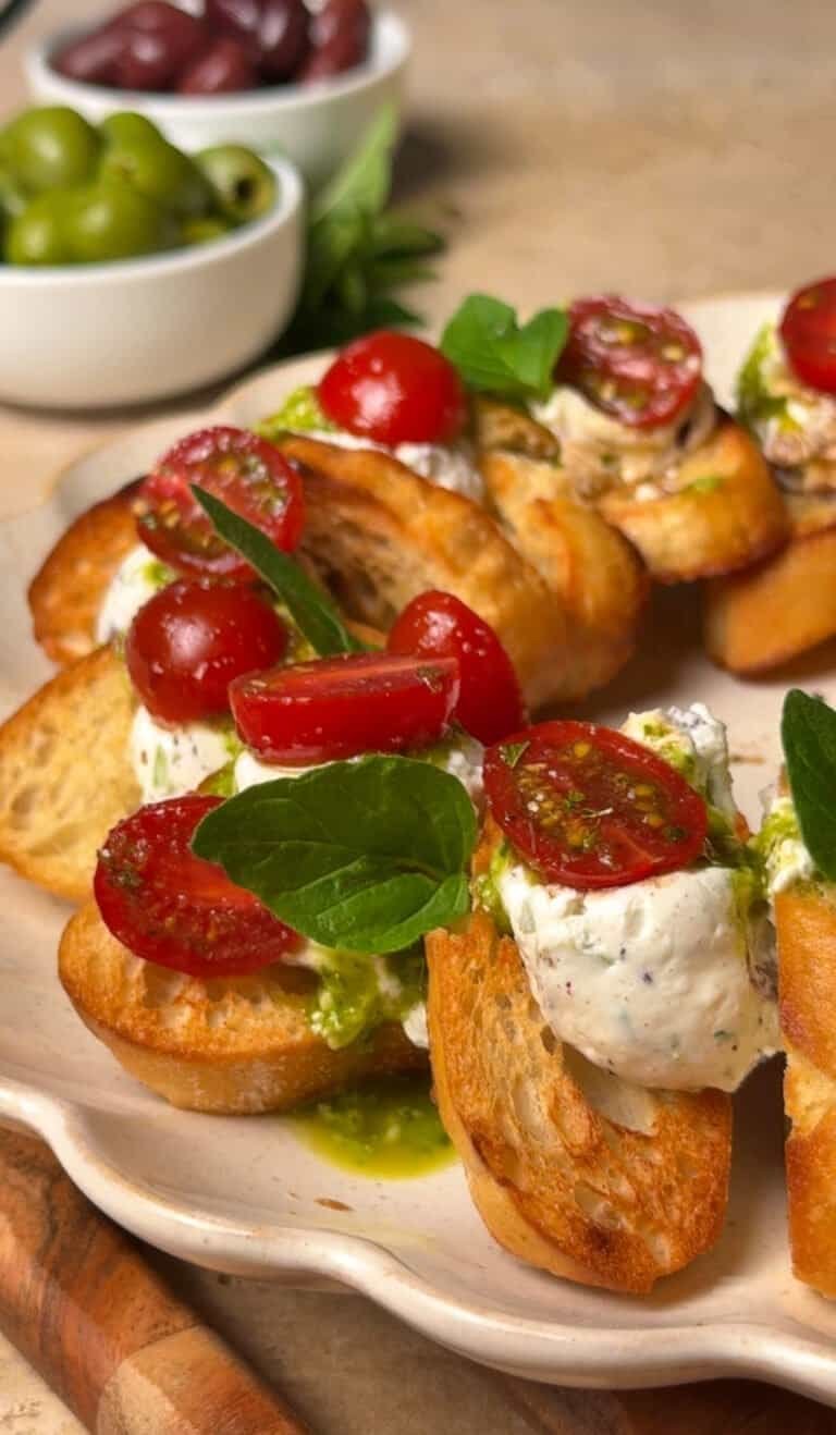 Close-up of whipped labneh crostini arranged in a wreath on a scalloped plate, topped with halved cherry tomatoes and herb pesto