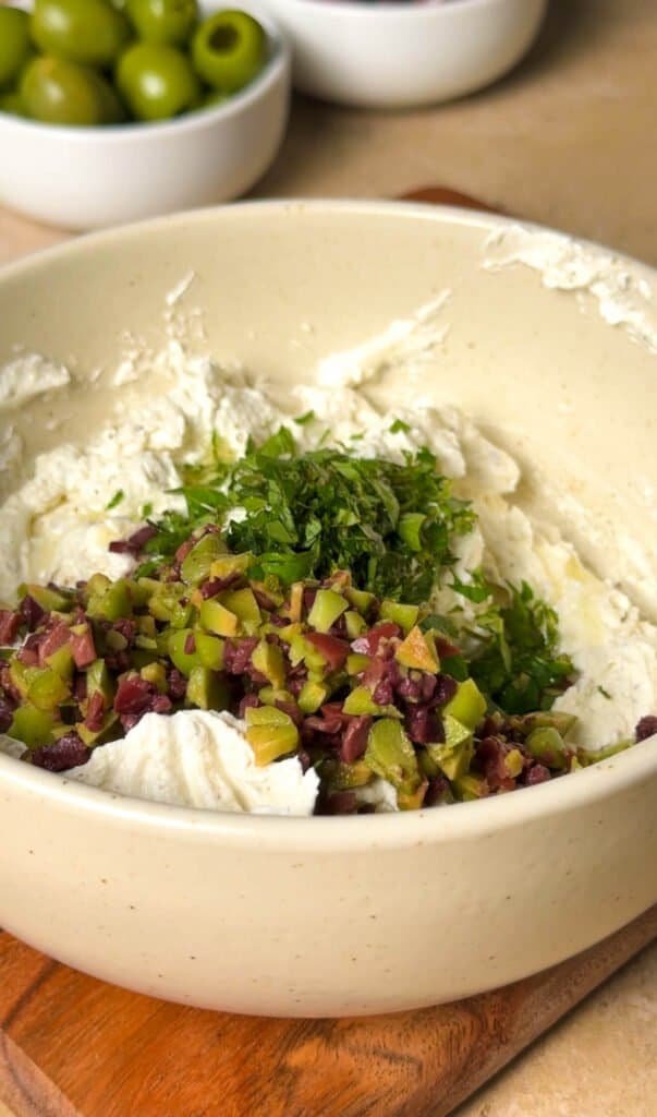Creamy labneh and French feta mixture in a ceramic bowl with finely chopped Castelvetrano and Kalamata olives and fresh oregano, before mixing