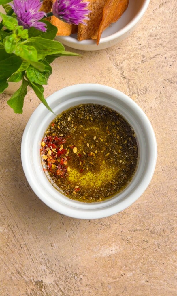 Za'atar olive oil with chili flakes in a small white bowl, with chive blossoms and pita chips in the background