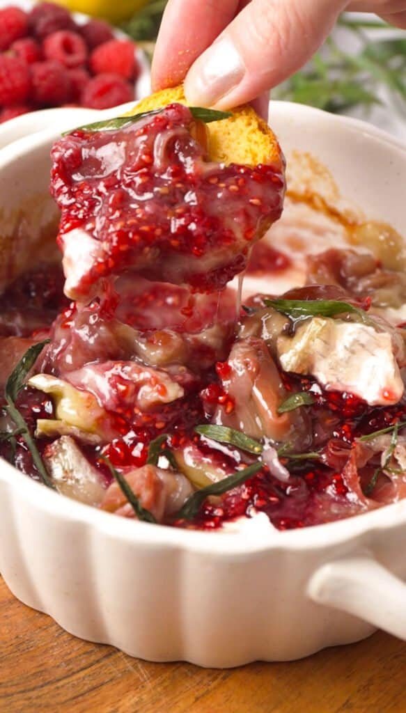 Crostini being pulled from baked brie with raspberry preserve, melted cheese stretching from the dish