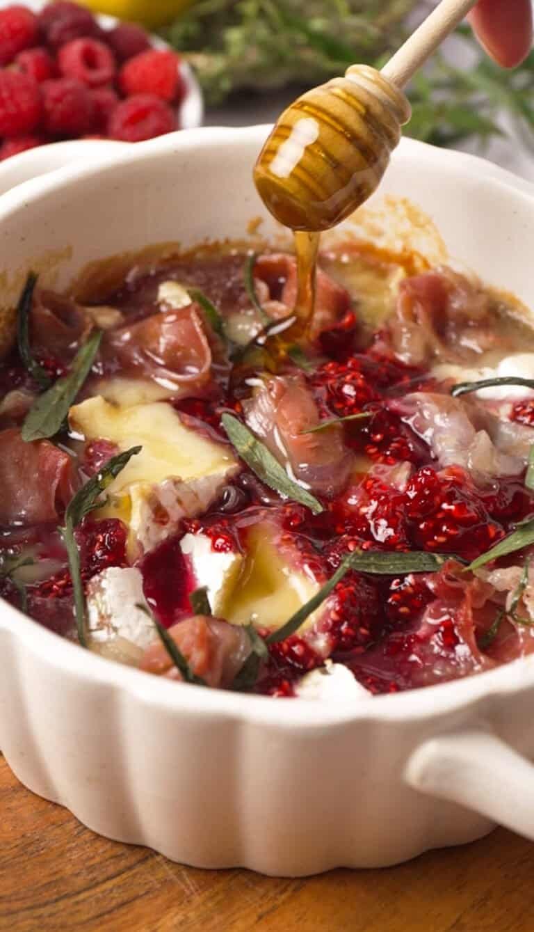 Hot honey being drizzled over baked brie with raspberry preserve and prosciutto in a white ceramic dish