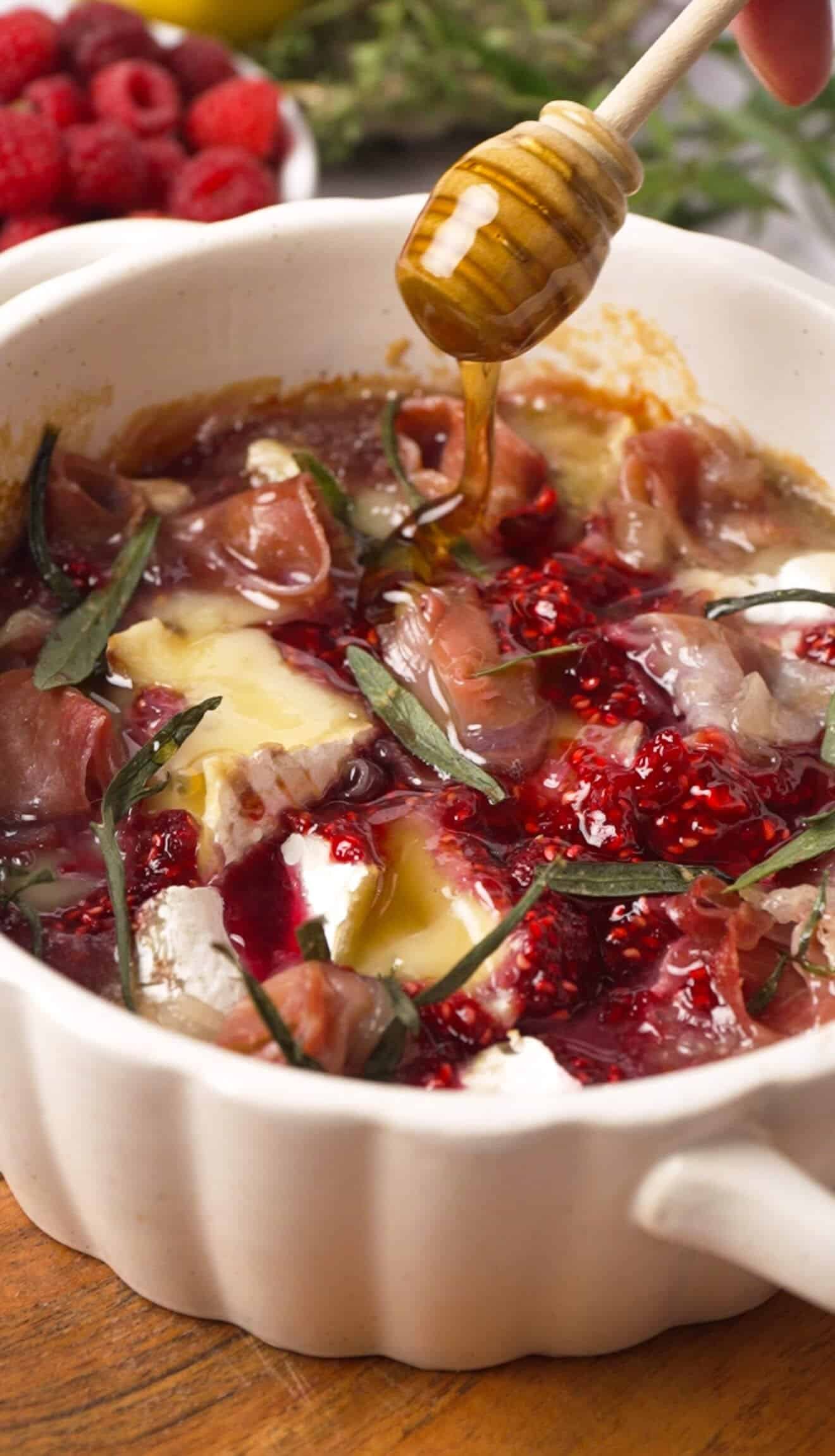 Hot honey being drizzled over baked brie with raspberry preserve and prosciutto in a white ceramic dish