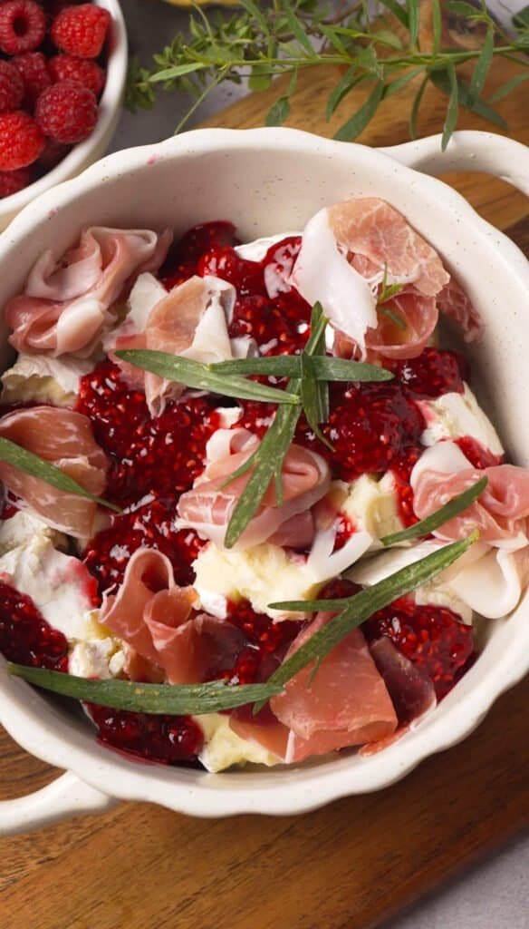 Baked brie with raspberry preserve, prosciutto, and fresh tarragon in a white ceramic dish before baking